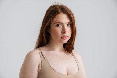 Duvarın arkasında durup kameraya bakan uzun kızıl saçlı, kıvrımlı genç bir kadının portresi.