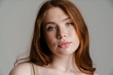 Duvarın arkasında durup kameraya bakan uzun kızıl saçlı, kıvrımlı genç bir kadının portresi.