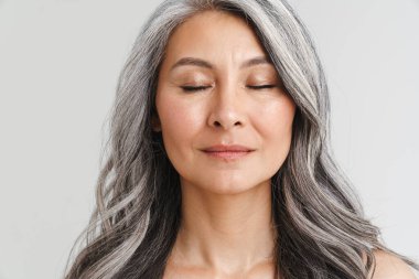 Olgun, üstsüz, gözleri kapalı poz veren bir kadın beyaz arka planda izole edilmiş kameraya bakıyor.