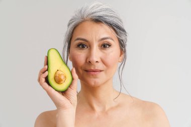 Beyaz arka planda avakado gösteren gri saçlı, olgun, üstsüz bir kadın.