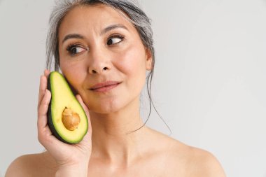 Beyaz arka planda avakado gösteren gri saçlı, olgun, üstsüz bir kadın.