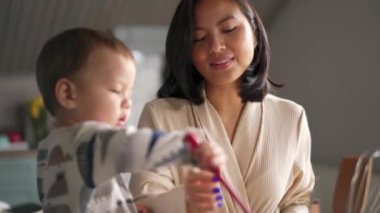 Gülümseyen Asyalı anne bebeğiyle konuşuyor. Mutfak spatulasıyla oynuyor.