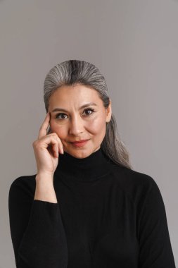 Beyaz saçlı olgun bir kadın. Gri arka planda yalnız başına poz veriyor ve kameraya bakıyor.