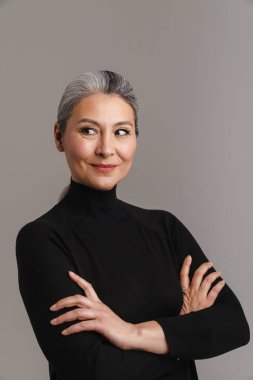 Olgun, beyaz saçlı, gülümseyen bir kadın kollarını kavuşturup gri arka planda izole bir şekilde poz veriyor.