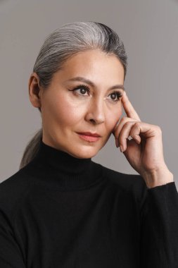 Beyaz saçlı olgun bir kadın. Gri arka planda yalnız başına poz veriyor ve bakıyor.