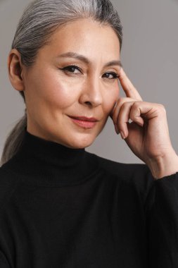 Beyaz saçlı olgun bir kadın. Gri arka planda yalnız başına poz veriyor ve kameraya bakıyor.