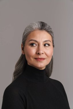 Beyaz saçlı olgun bir kadın. Gri arka planda yalnız başına poz veriyor ve bakıyor.