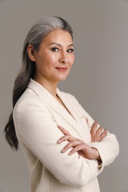 Olgun Asyalı beyaz saçlı kadın kollarını kavuşturup gri arka planda izole bir şekilde poz veriyor.