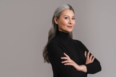 Olgun Asyalı beyaz saçlı kadın kollarını kavuşturup gri arka planda izole bir şekilde poz veriyor.