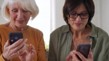 Neşeli orta yaşlı kadınlar evdeki kanepede telefonla mesajlaşıyorlar.