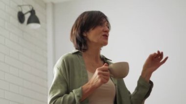 Kendinden emin, olgun bir kadın mutfakta elinde bardakla konuşuyor.