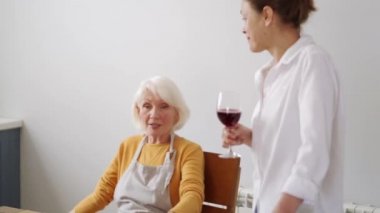 Gülümseyen olgun kadınlar ellerinde şarap, mutfakta konuşuyorlar.