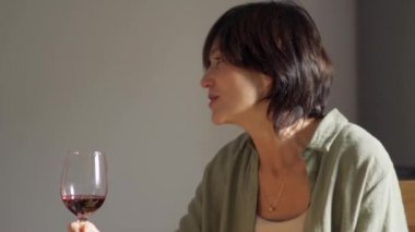 Konsantre esmer, olgun bir kadın mutfakta bir bardak şarapla konuşuyor.