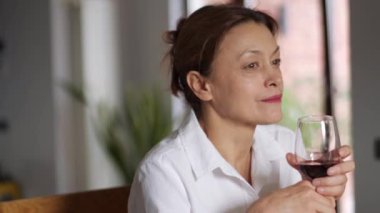 Düşünceli, olgun bir kadın mutfakta bir bardak şarapla arkadaşlarını dinliyor.