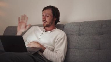 Evdeki kanepede otururken bilgisayarla konuşan uzun saçlı yakışıklı adam.