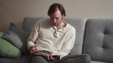 Evde kitap okuyan dalgın, uzun saçlı bir adam.
