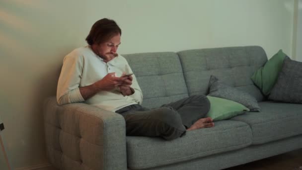 一个严肃的长发男人坐在沙发上 一边打电话一边发短信 — 图库视频影像