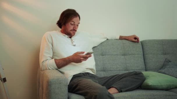 Skoncentrowany Długowłosy Mężczyzna Smsuje Przez Telefon Siedząc Kanapie Domu — Wideo stockowe