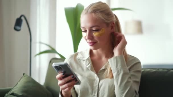 Poważna Blondynka Przyklejonymi Łatami Oczu Smsująca Przez Telefon Domu — Wideo stockowe