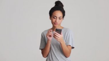 Tişörtlü neşeli Afrikalı kadın gri stüdyoda telefonla kayıt yapıyor.