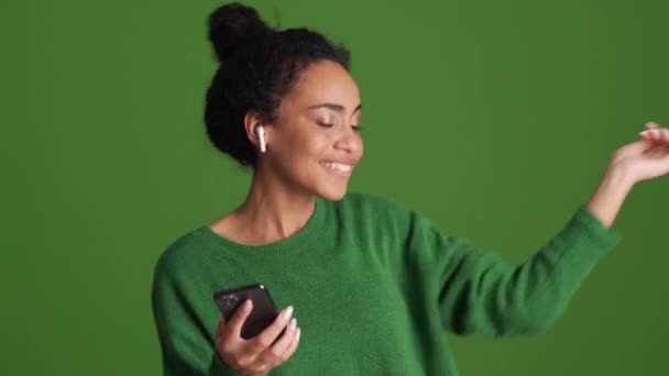 笑顔アフリカの女性で緑のシャツダンスと緑のスタジオでヘッドフォンで音楽を聴く — ストック動画