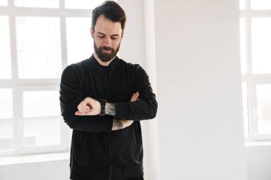 Sakin, esmer, spor kıyafetli beyaz bir adam. Kapalı mekanda dikilip akıllı bakışlara bakıyor.