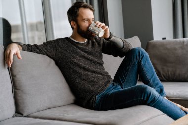 Sakallı Avrupalı adam evdeki kanepede otururken kahve içiyor.