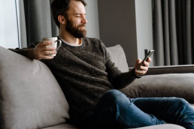 Sakallı adam evde kanepede otururken cep telefonu kullanıyor ve kahve içiyor.