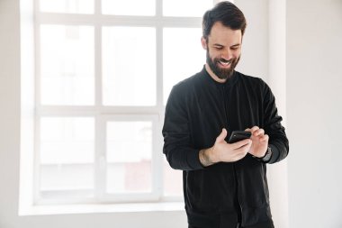 Gülümseyen esmer, beyaz bir adam spor giyim kuşamında evde durmuş cep telefonuna bakıyor.