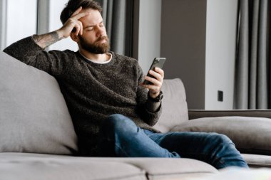 Sakallı Avrupalı adam evdeki koltukta otururken cep telefonu kullanıyor.