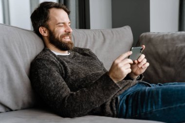 Sakallı Avrupalı adam evdeki koltukta otururken cep telefonu kullanıyor.