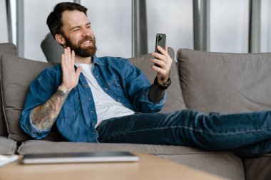 Sakallı Avrupalı adam evdeki kanepede otururken el kol hareketi yapıyor ve cep telefonu kullanıyor.