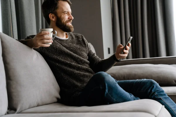 Homem Barbudo Usando Telefone Celular Beber Café Enquanto Sentado Sofá — Fotografia de Stock