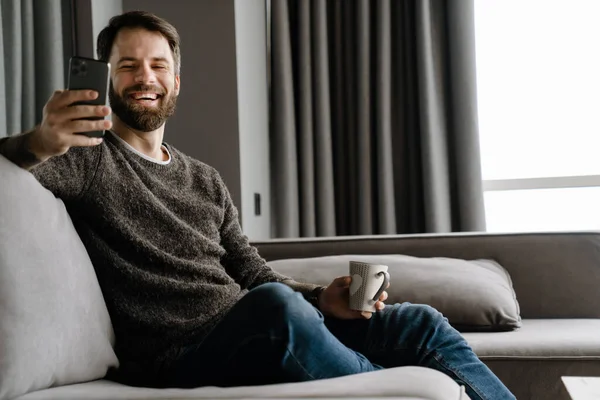 Bearded Man Using Mobile Phone Drinking Coffee While Sitting Sofa — Stock Photo, Image