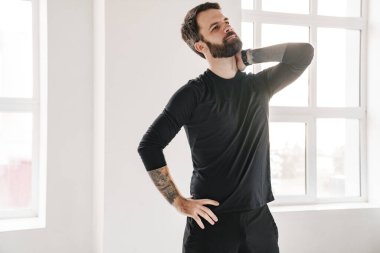 Sakin, orta yaşlı, sakallı, spor kıyafetli bir adam. Pencere kenarında durup, boynunu ovalıyor.