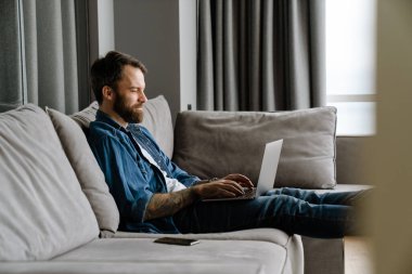 Sakallı Avrupalı adam evdeki kanepede otururken dizüstü bilgisayarla çalışıyor.