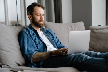 Sakallı Avrupalı adam evdeki kanepede otururken dizüstü bilgisayarla çalışıyor.