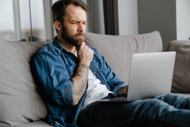 Sakallı Avrupalı adam evdeki kanepede otururken dizüstü bilgisayarla çalışıyor.