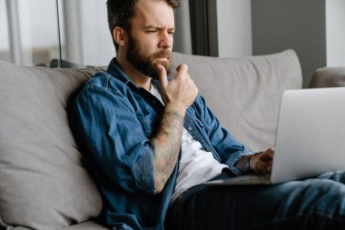 Sakallı Avrupalı adam evdeki kanepede otururken dizüstü bilgisayarla çalışıyor.
