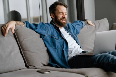 Sakallı Avrupalı adam evdeki kanepede otururken dizüstü bilgisayarla çalışıyor.