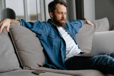 Sakallı Avrupalı adam evdeki kanepede otururken dizüstü bilgisayarla çalışıyor.