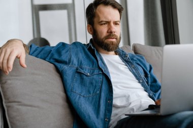 Sakallı Avrupalı adam evdeki kanepede otururken dizüstü bilgisayarla çalışıyor.