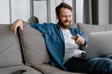 Sakallı adam evdeki kanepede otururken dizüstü bilgisayarla hareket ediyor ve çalışıyor.