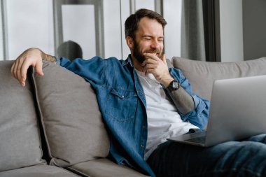 Sakallı adam evde kanepede otururken bilgisayarla gülüp çalışıyor.