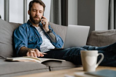 Sakallı adam evde çalışırken cep telefonuyla konuşuyor ve laptop kullanıyor.