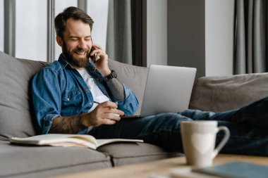 Sakallı adam evde çalışırken cep telefonuyla konuşuyor ve laptop kullanıyor.