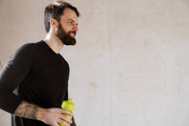 Orta yaşlı, gülümseyen, sakallı, elinde su şişesi olan beyaz bir adam.