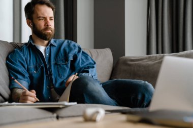 Sakallı Avrupalı adam evdeki kanepede otururken notlar yazıyor.