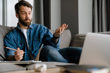 Sakallı Avrupalı adam evdeki kanepede otururken dizüstü bilgisayarla hareket ediyor ve çalışıyor.