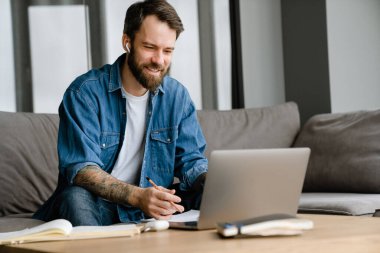 Sakallı Avrupalı adam evdeki kanepede otururken dizüstü bilgisayarla çalışıyor.